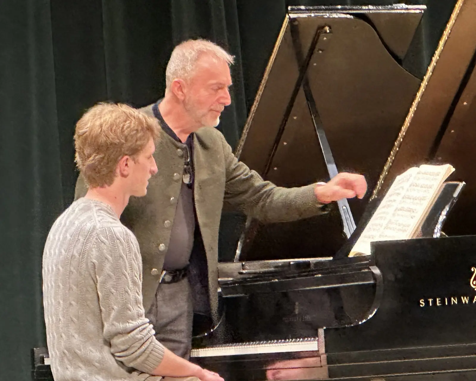 An instructor points to sheet music for a student seated at a grand piano.