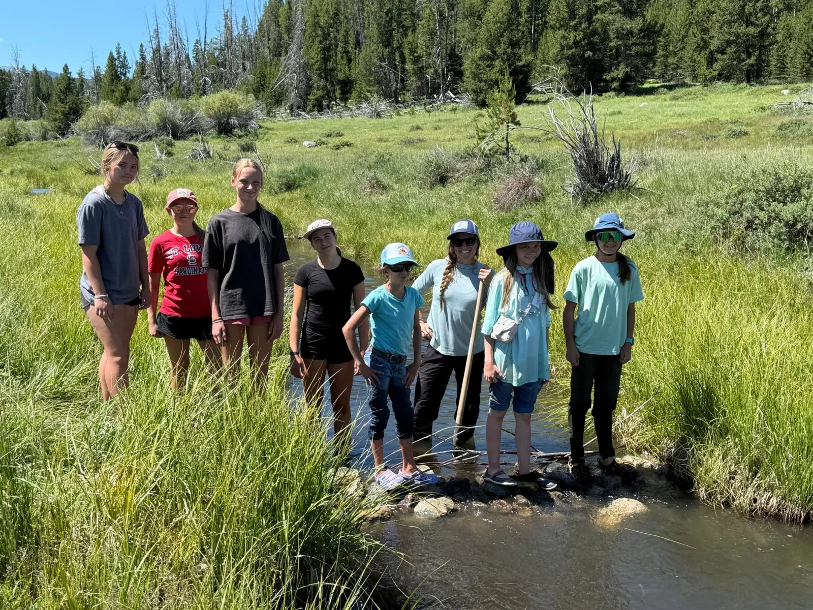 A group of young people and an adult stand together in a shallow stream during an outdoor excursion.