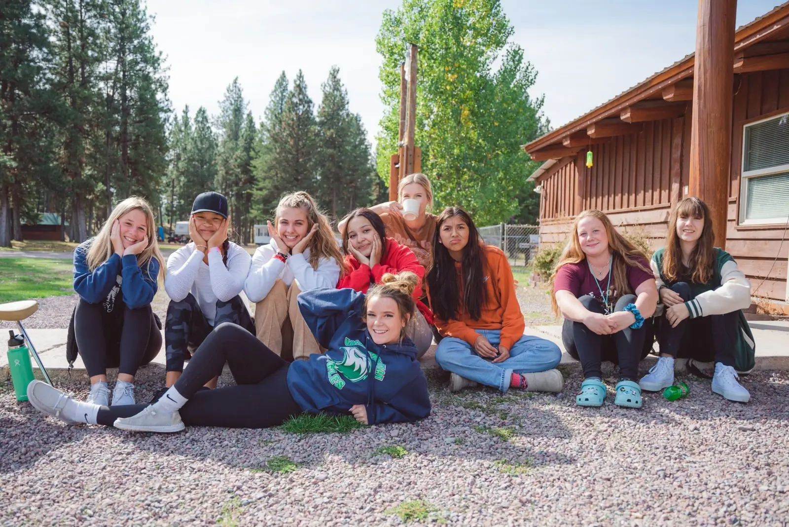 A group of young women pose for a photograph while sitting on the ground outside a wooden building.