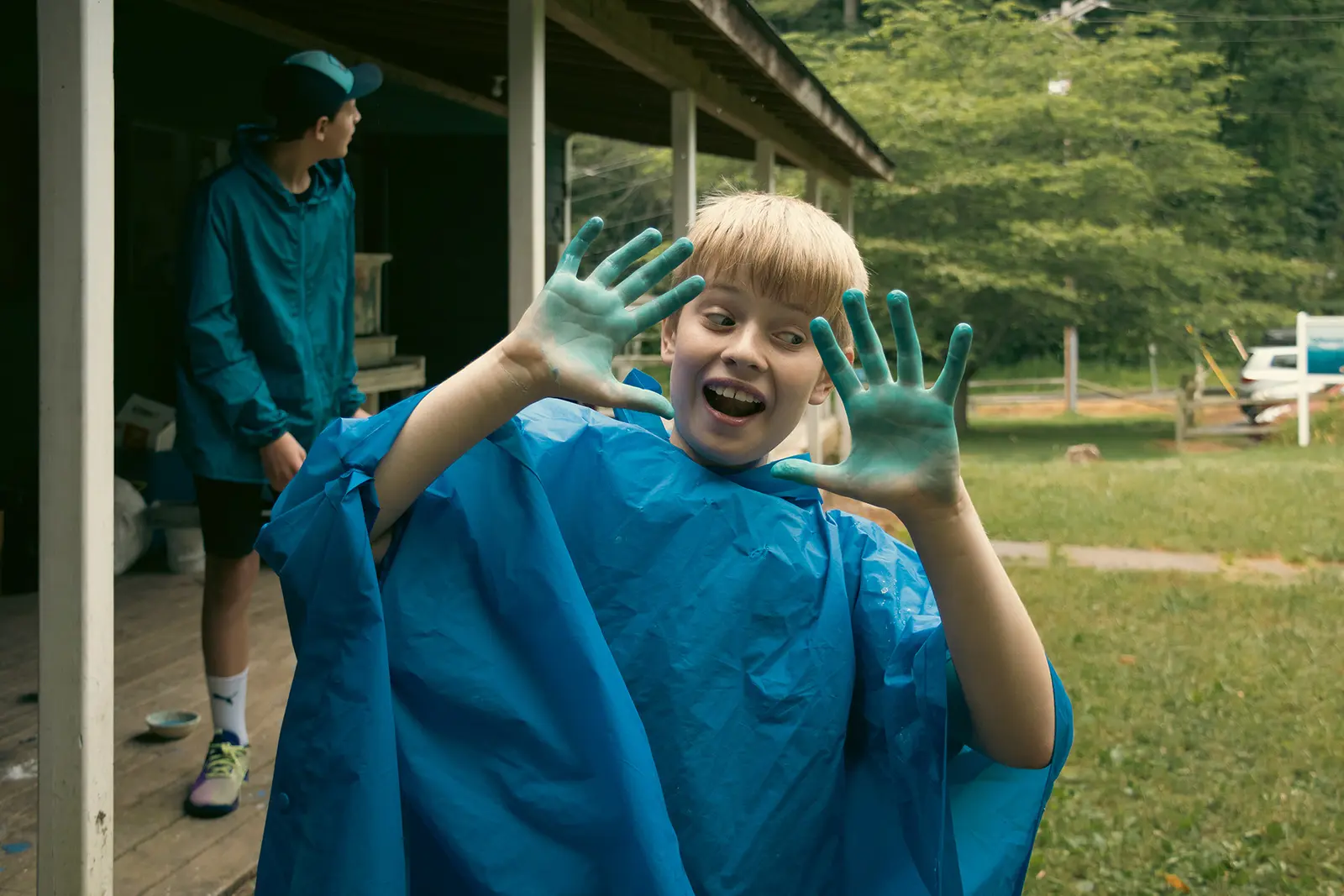 A boy with blue-painted hands and a blue poncho smiles excitedly outdoors.