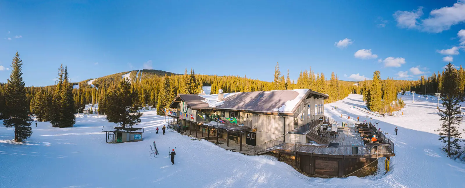 A panoramic view shows a ski lodge surrounded by snow and evergreen trees on a sunny day.