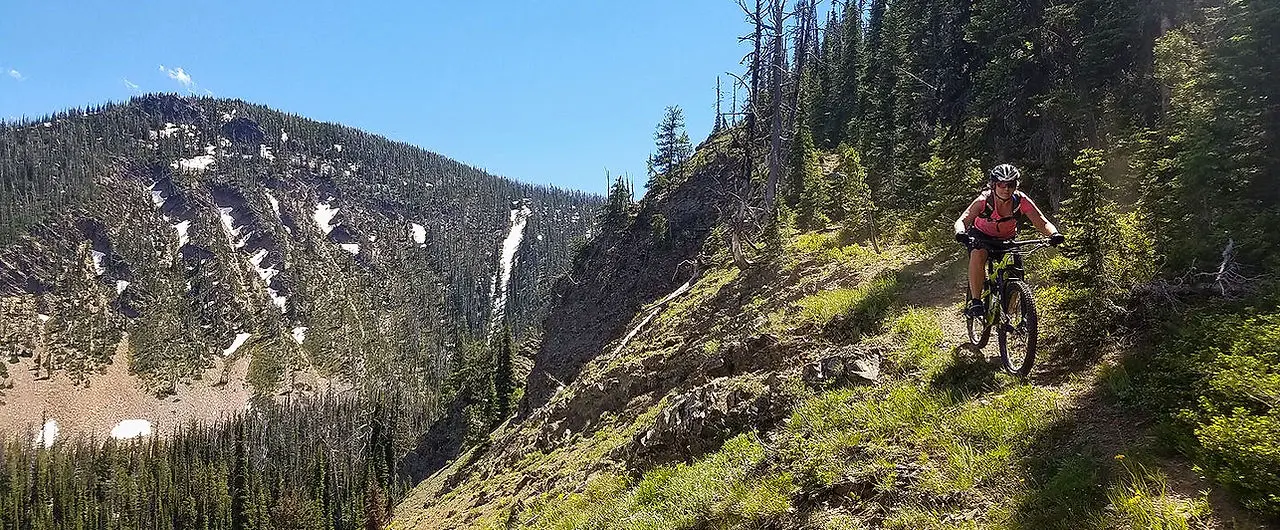 A person rides a mountain bike on a sunny trail with mountains in the background.