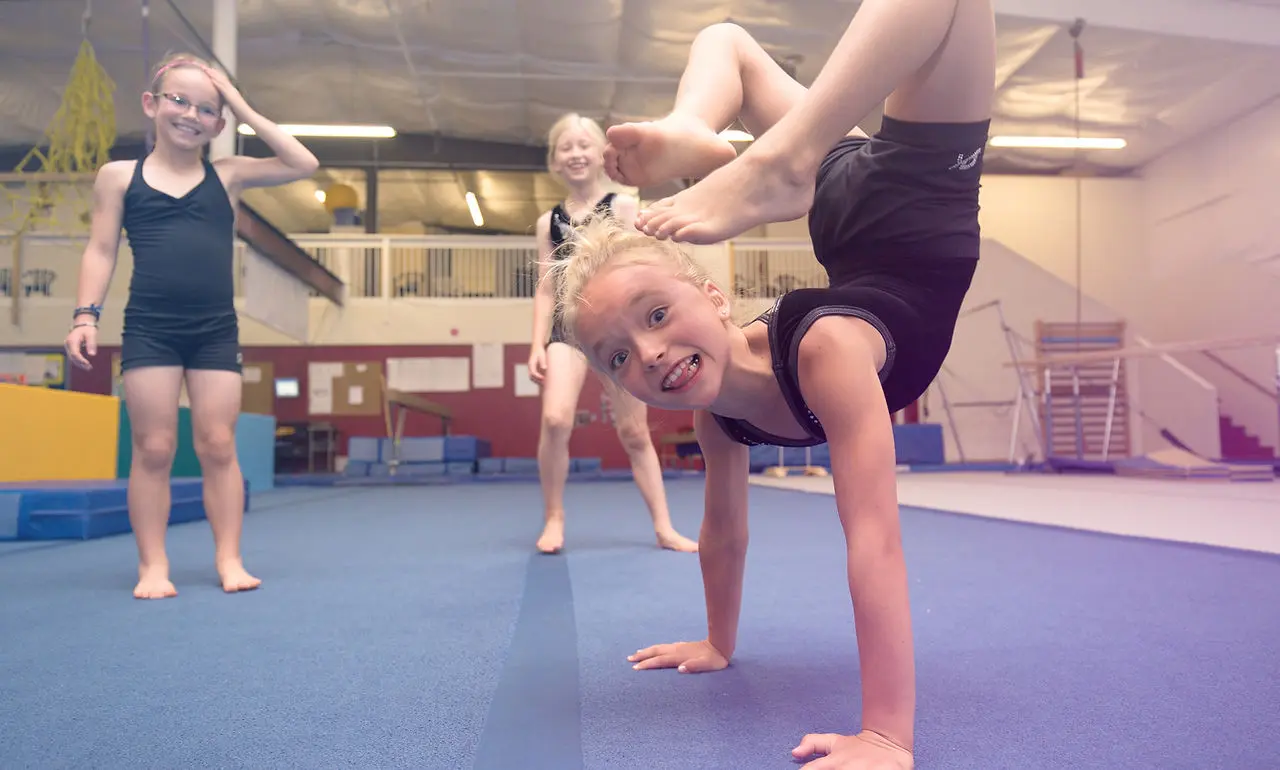 Three girls in athletic wear are in a gymnastics gym with one performing a handstand.