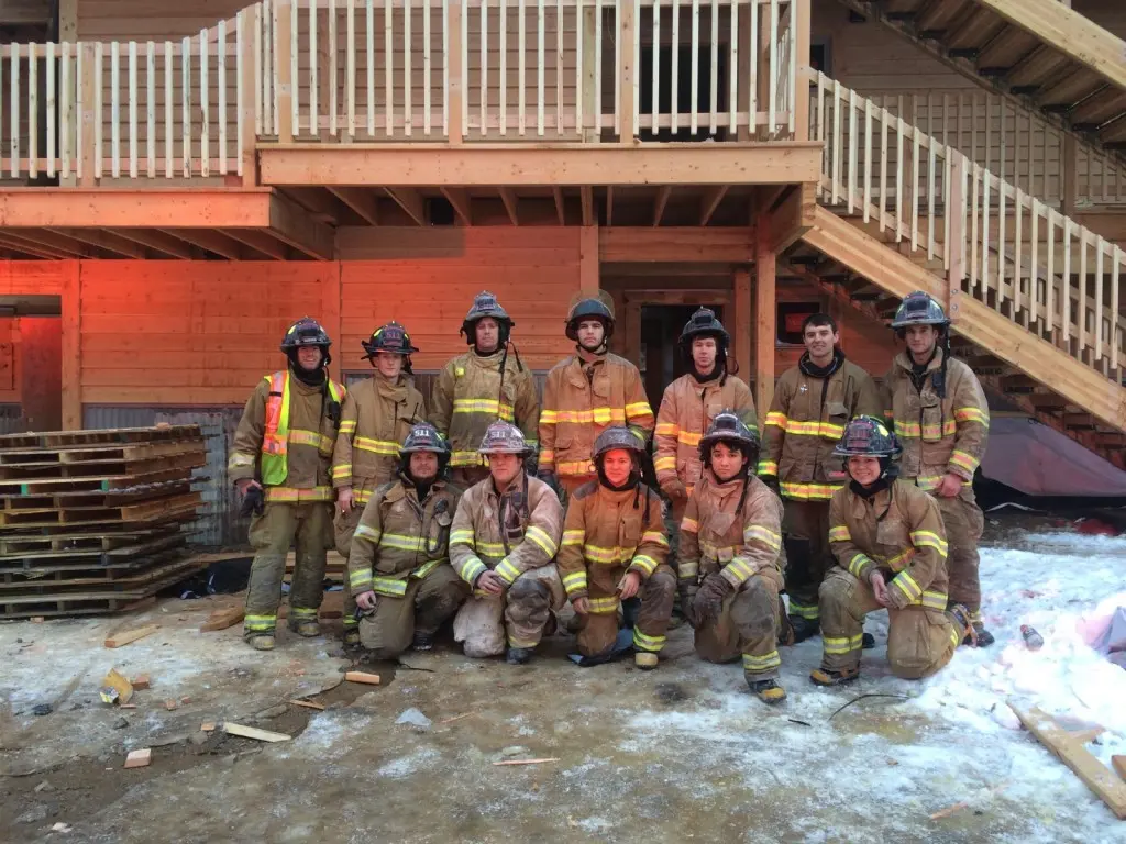 A group of firefighters in gear pose for a photo in front of a wooden building under construction.