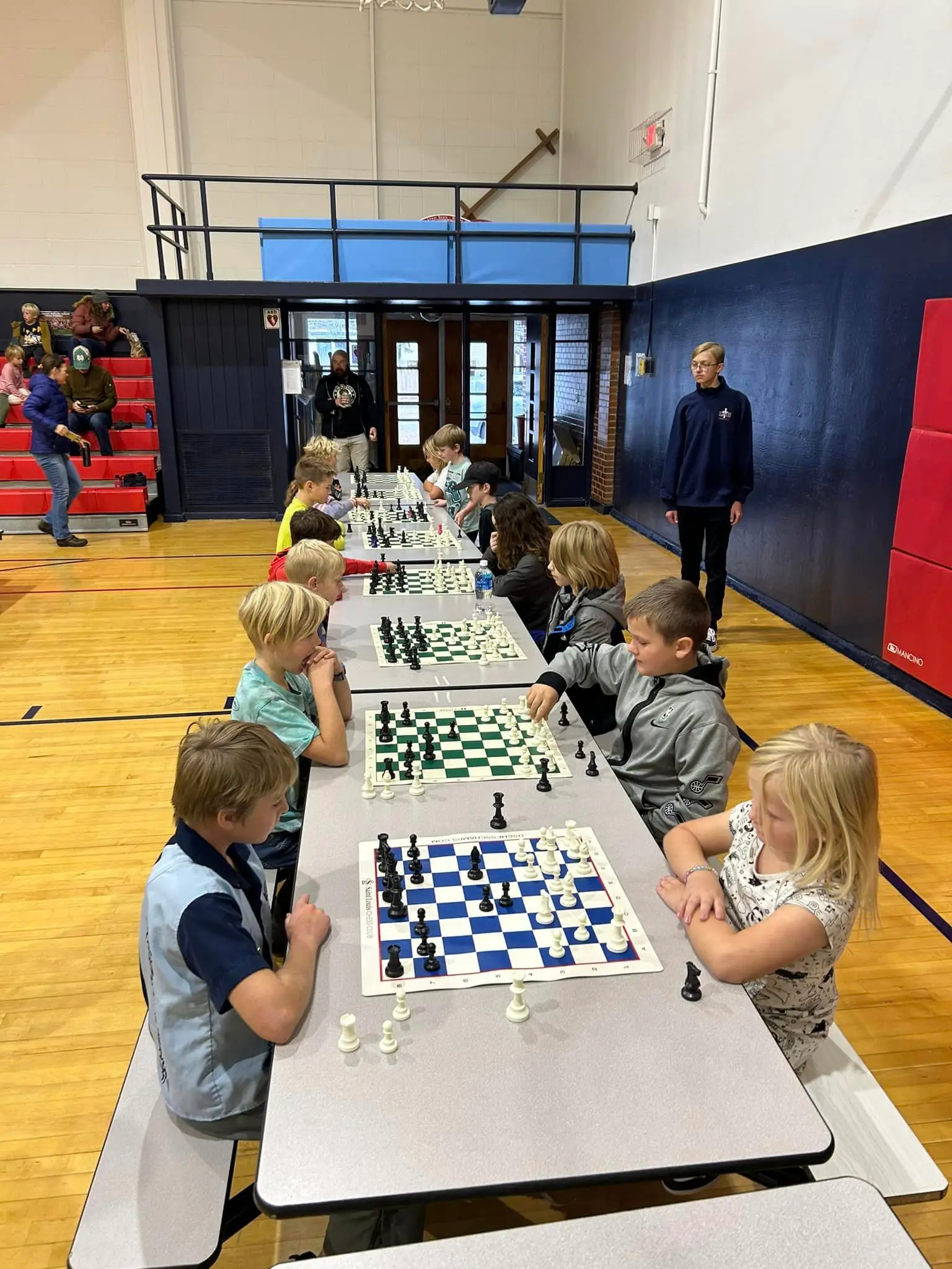 A group of children are sitting at tables in a gymnasium playing chess.