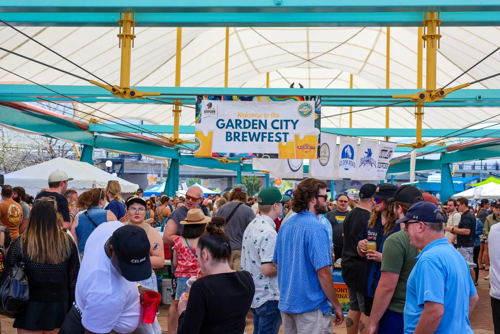 A crowd of adults enjoys an outdoor festival under a large canopy featuring a banner for a beer festival.