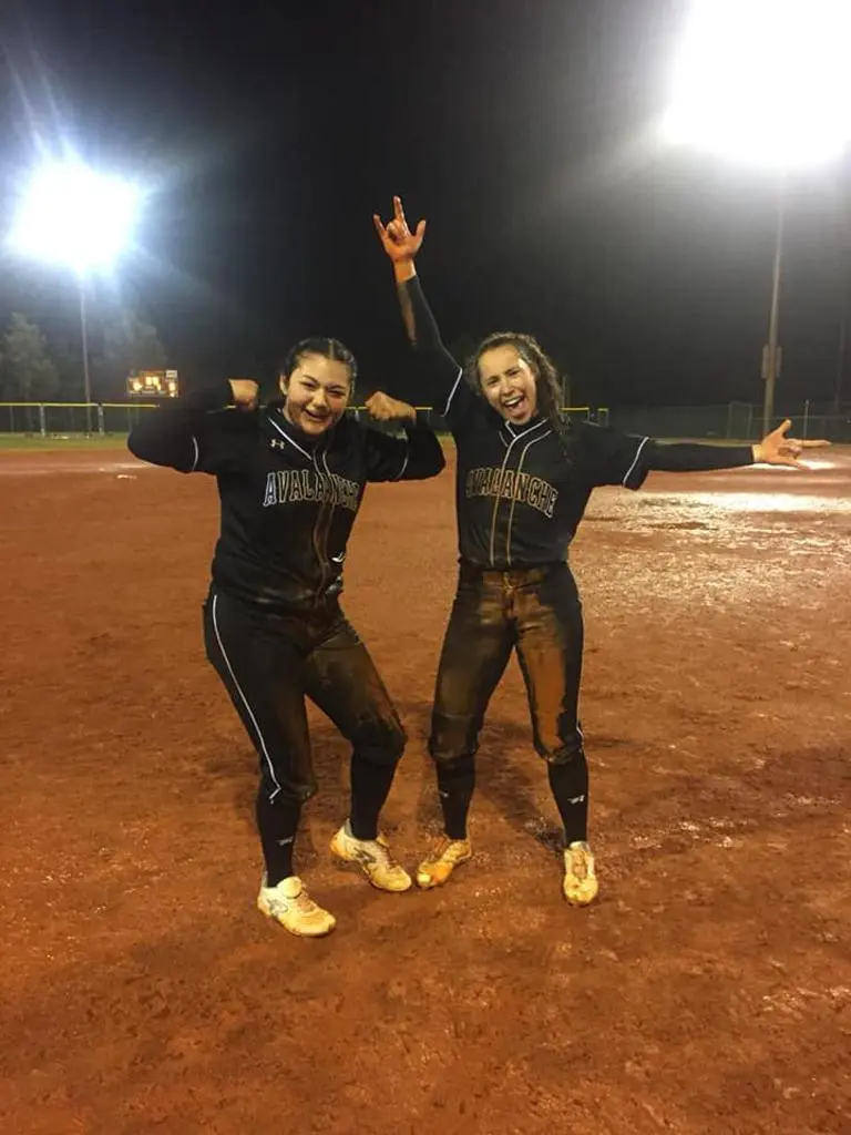 Two softball players in team uniforms pose on a field at night.