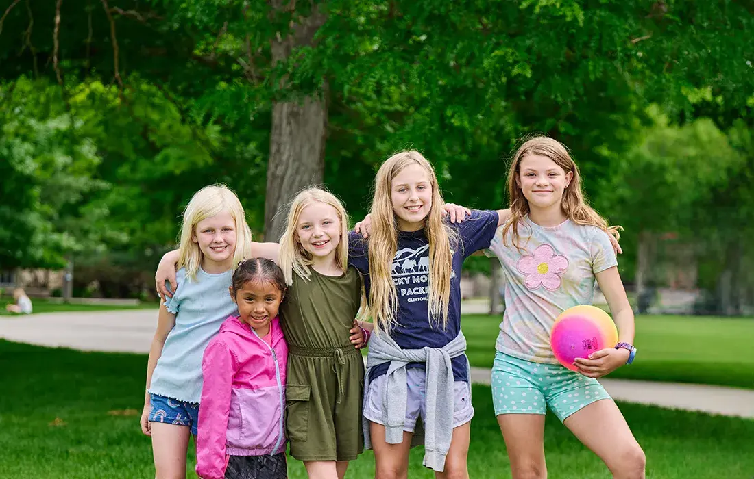 A group of five young girls stands smiling together in a grassy park setting.