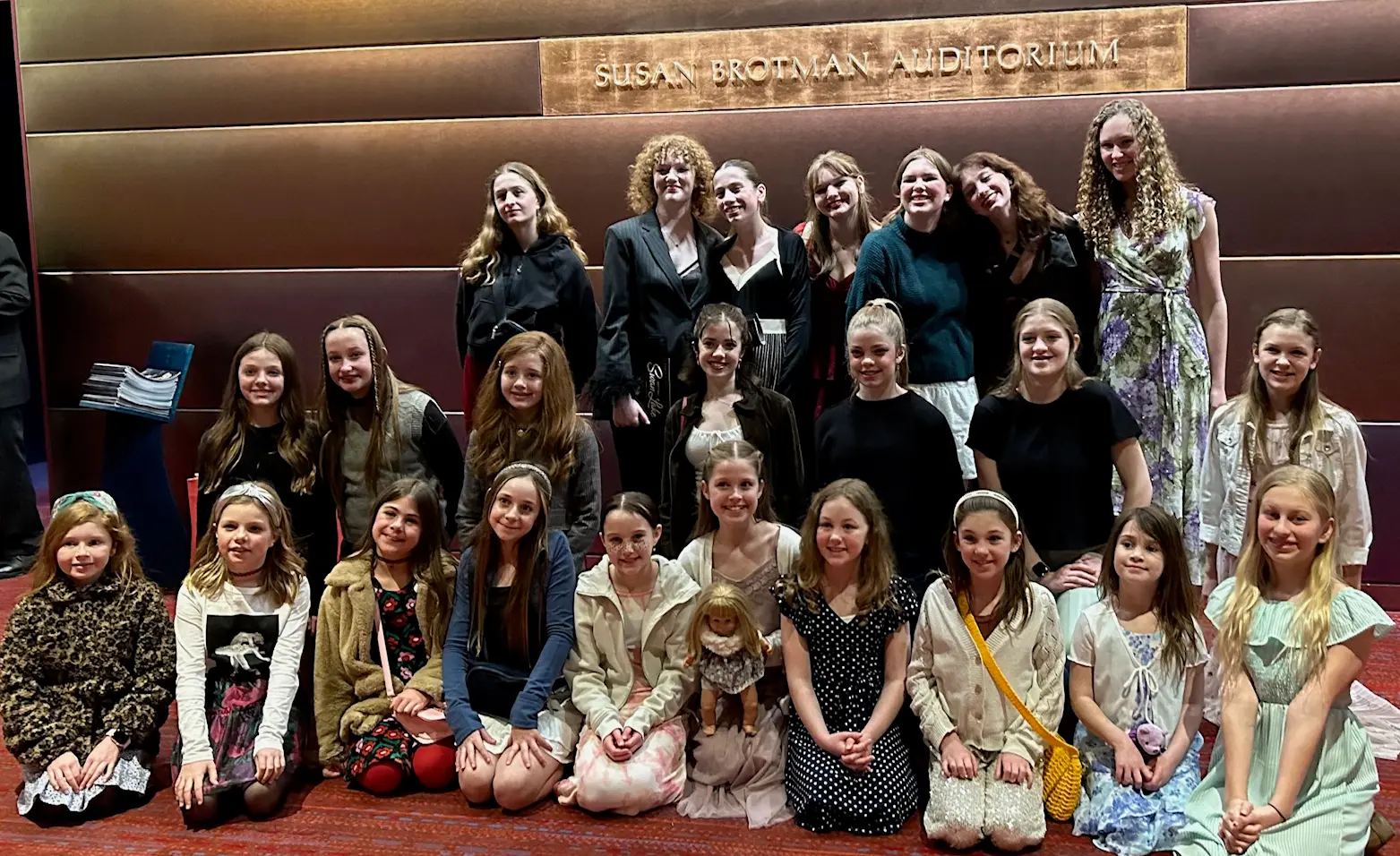 A large group of girls and young women pose for a photograph in front of an auditorium sign.