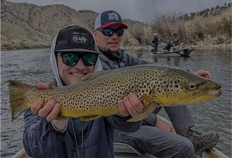Two men hold a large trout while sitting in a boat on a river.