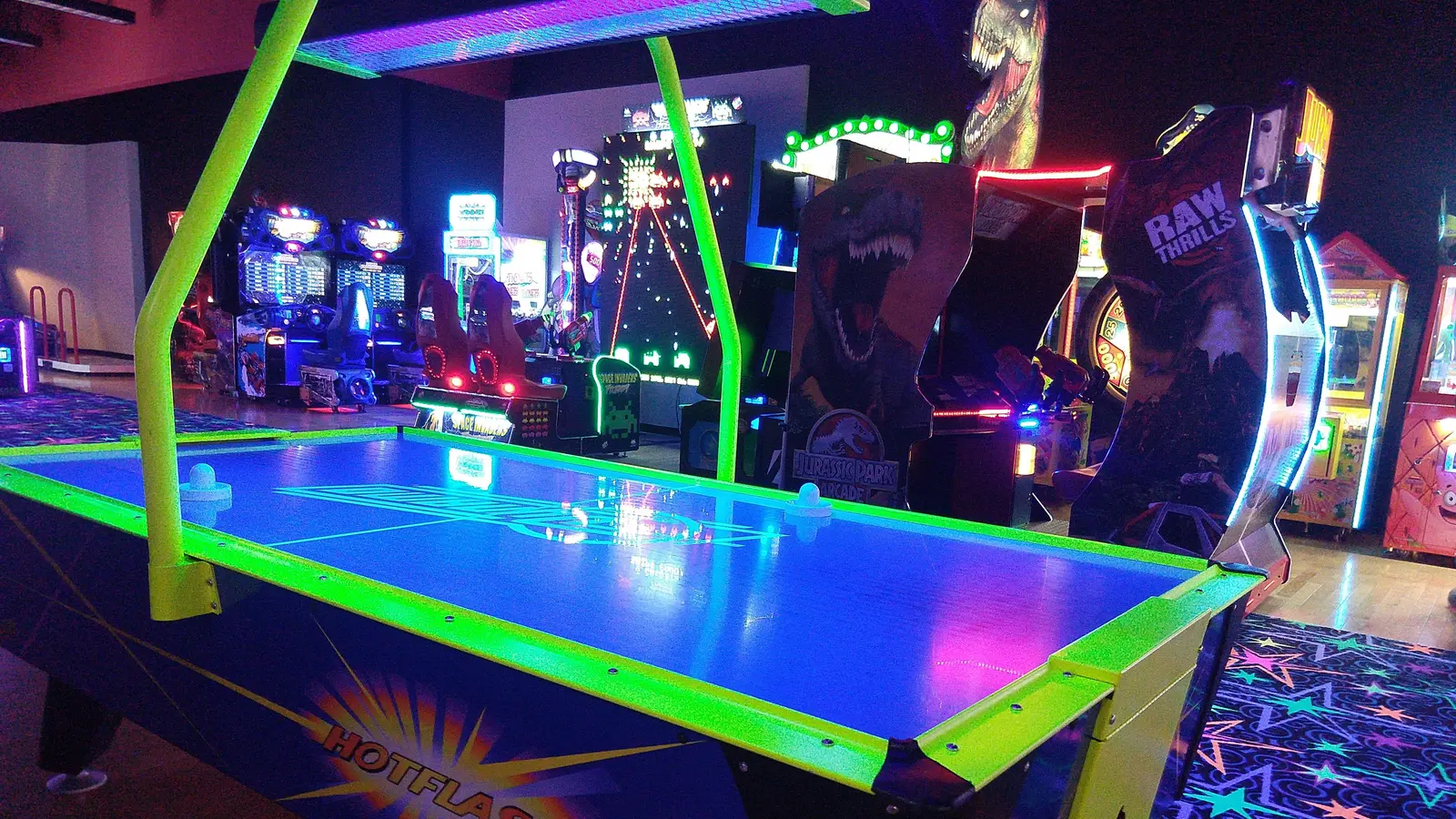 An indoor arcade features an air hockey table in the foreground and various video games in the background.