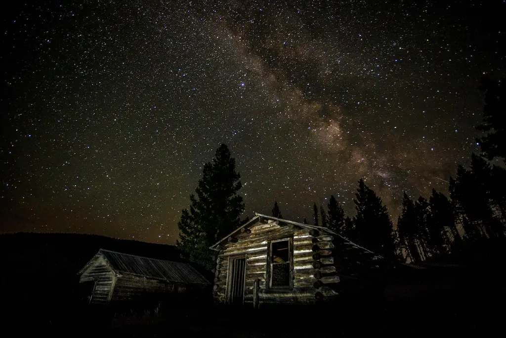 A rustic log cabin sits under a starry night sky with the Milky Way visible.