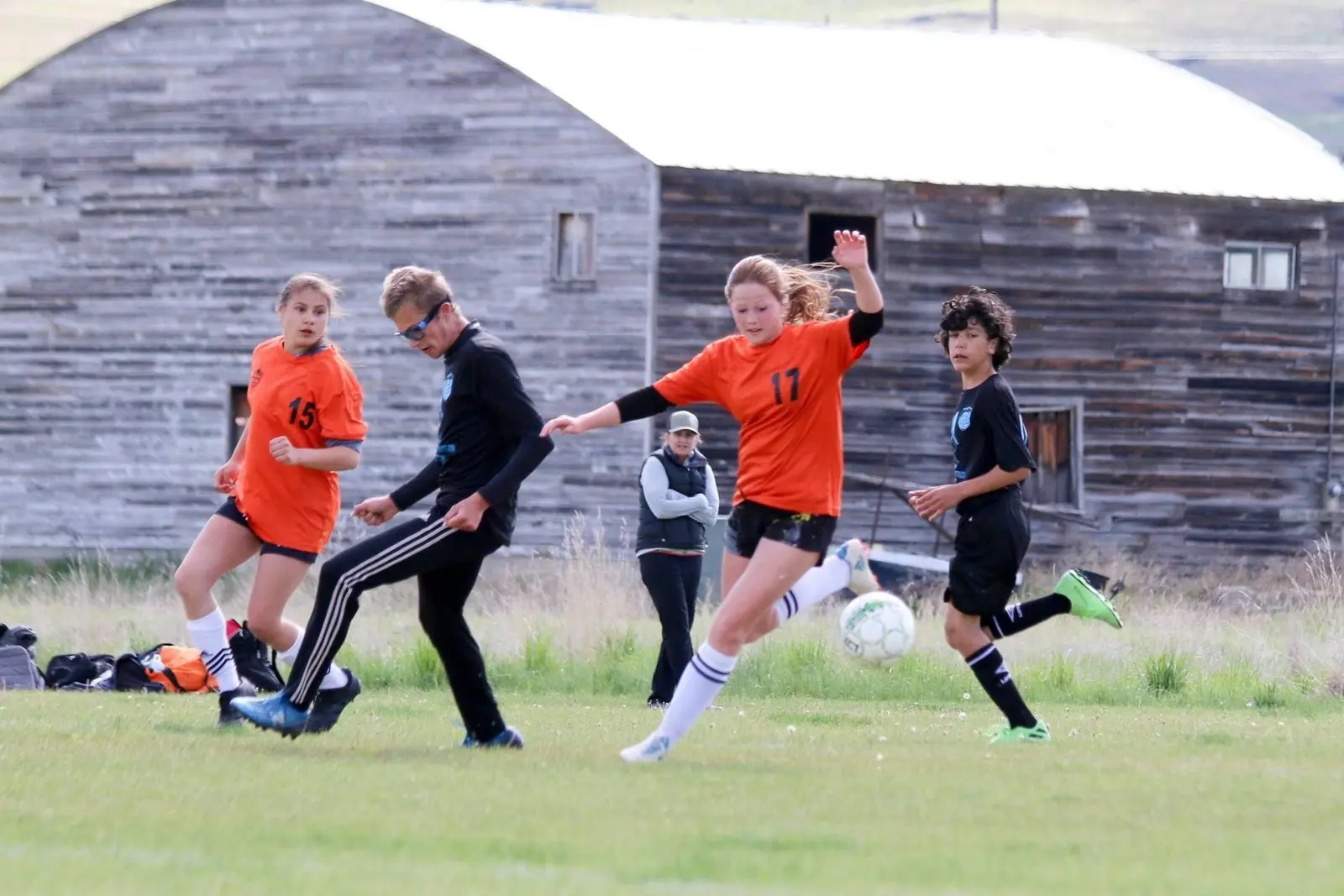 A group of young people play a game of soccer on a field in front of a rustic wooden building.