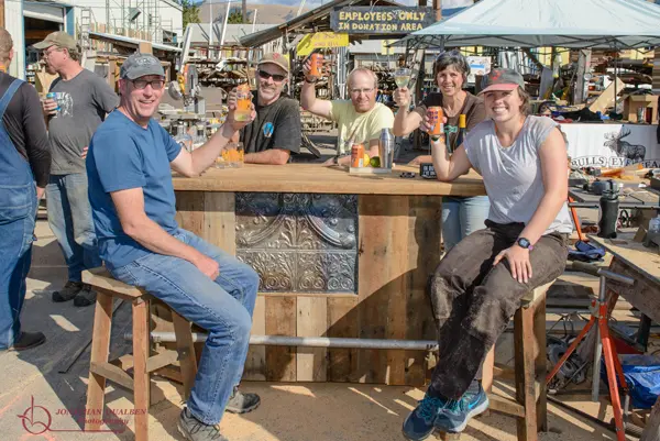 A group of adults are gathered around a makeshift wooden bar in what appears to be a workshop or yard.