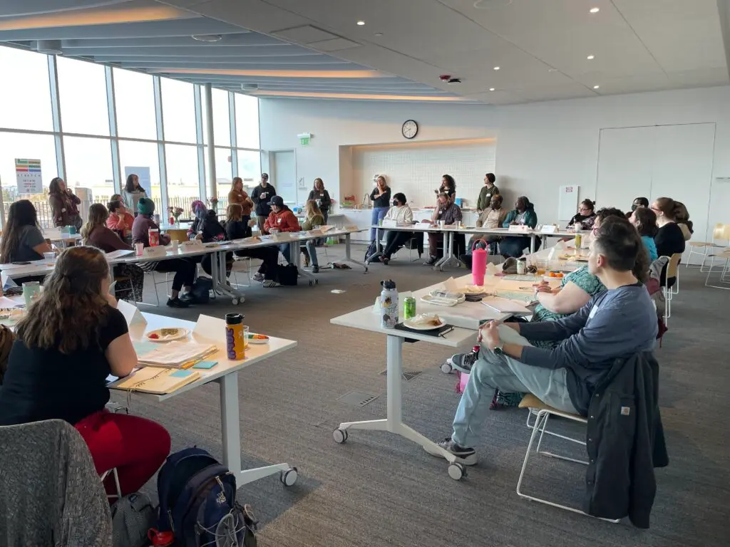 A group of adults sits at tables in a large, sunlit meeting room for a workshop or seminar.