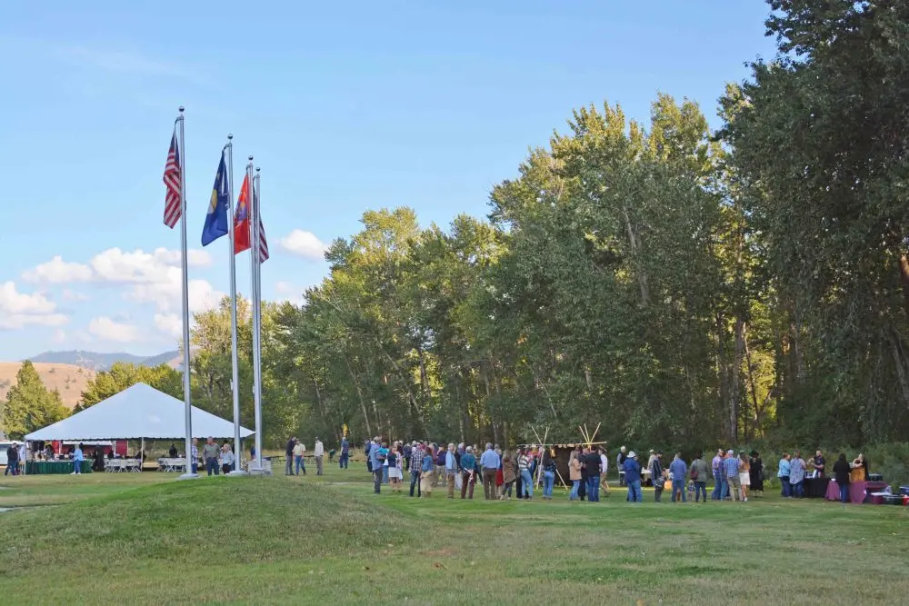 A crowd of people gathers in a grassy outdoor field near flagpoles and a white tent.