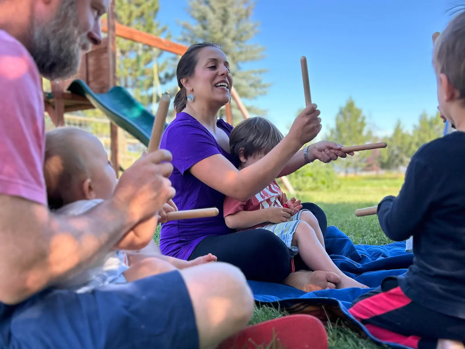 A family sits together on a blanket outdoors while holding wooden sticks.