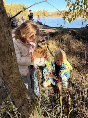 Two children are outdoors by a river, one drawing in a sketchbook while the other inspects something on the ground.