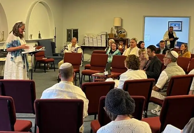 A group of adults sits in a room with rows of chairs while a person stands at the front reading from a book.