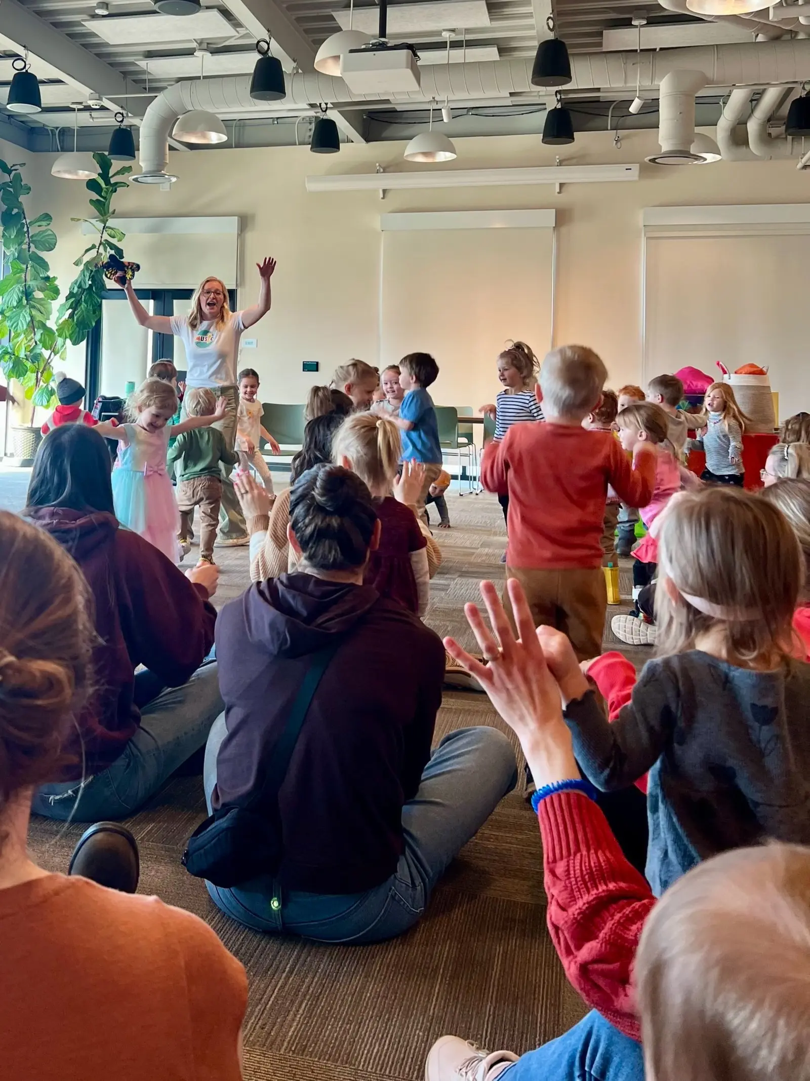 Children participating in a dance or activity class led by an instructor with long blonde hair, wearing glasses and a white T-shirt, inside a modern room with a ceiling of exposed pipes and large windows with blinds.