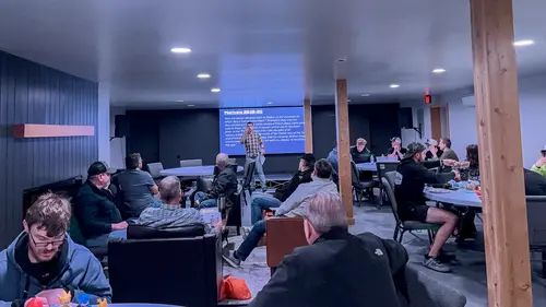 A presenter stands in front of a room of adults seated at tables for a presentation.