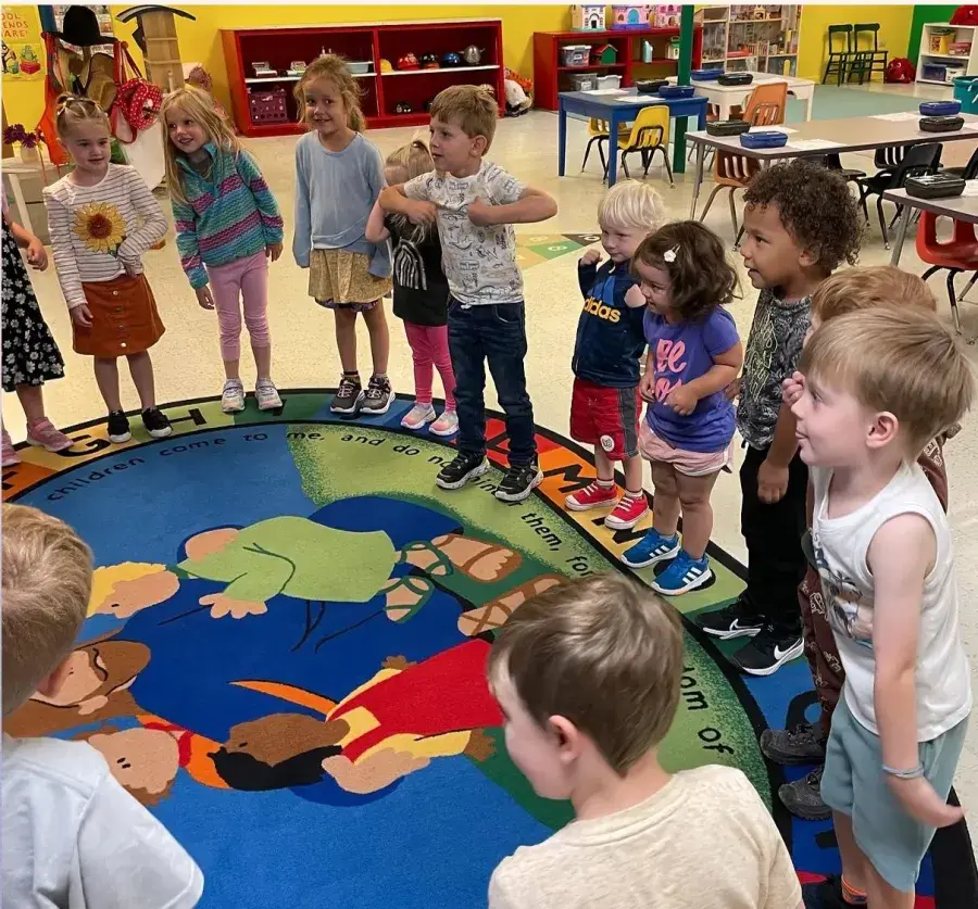 A group of young children stands in a circle on a colorful rug inside a classroom.