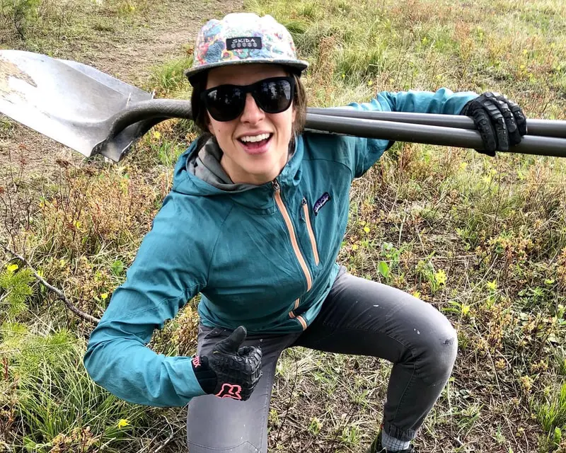 A person in outdoor gear smiles and gives a thumbs up while holding a shovel across their shoulders in a field.