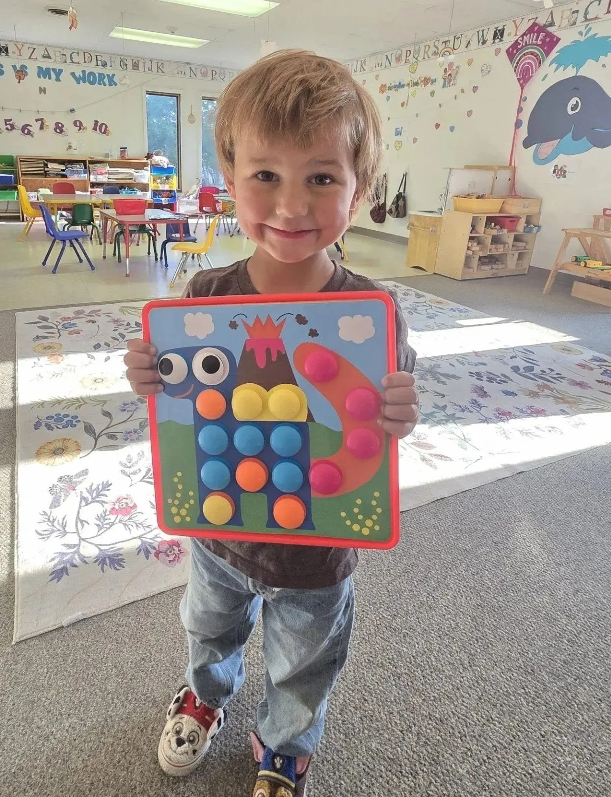 A young boy smiles while holding a dinosaur-shaped toy in a classroom setting.