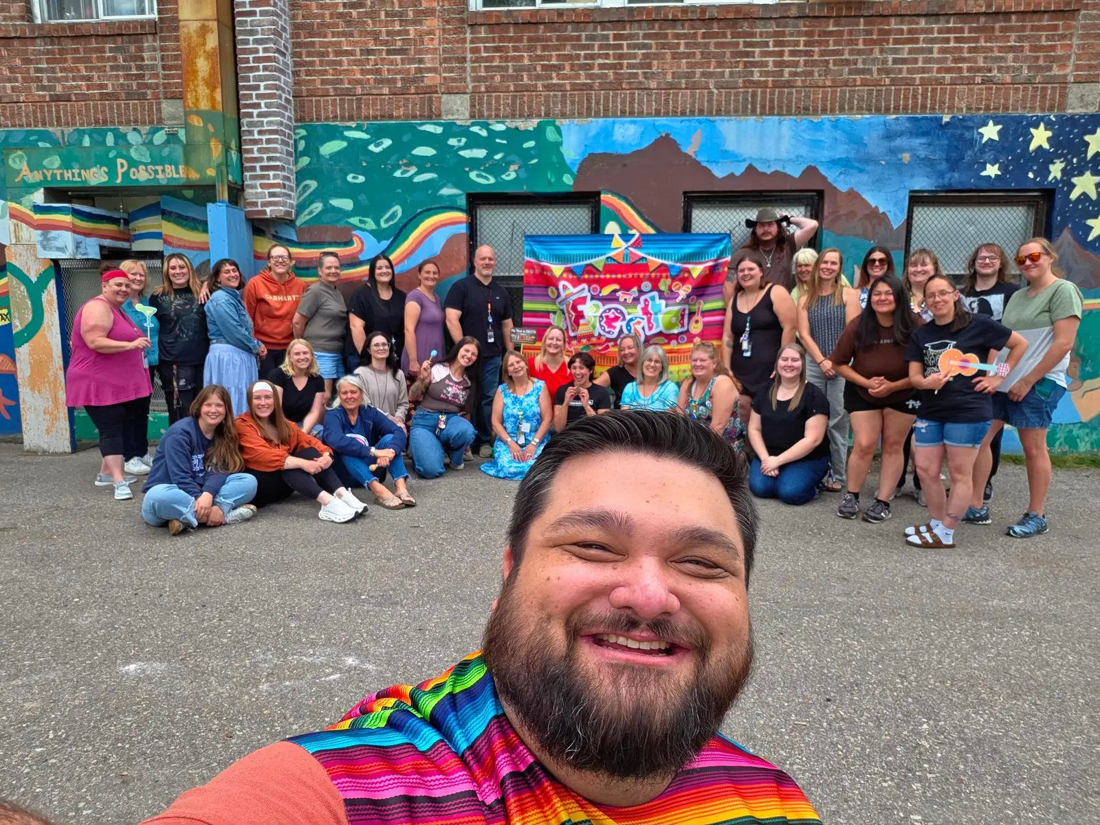 A large group of adults pose for a selfie in front of a brick wall with a mural and a festive banner.