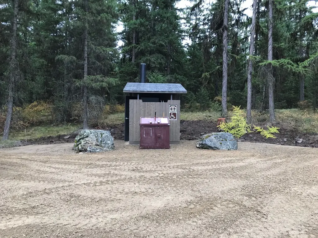 An outdoor vault toilet is situated in a clearing surrounded by a pine forest.