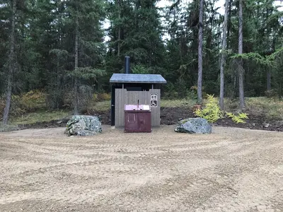 An outdoor vault toilet is situated in a clearing surrounded by a pine forest.