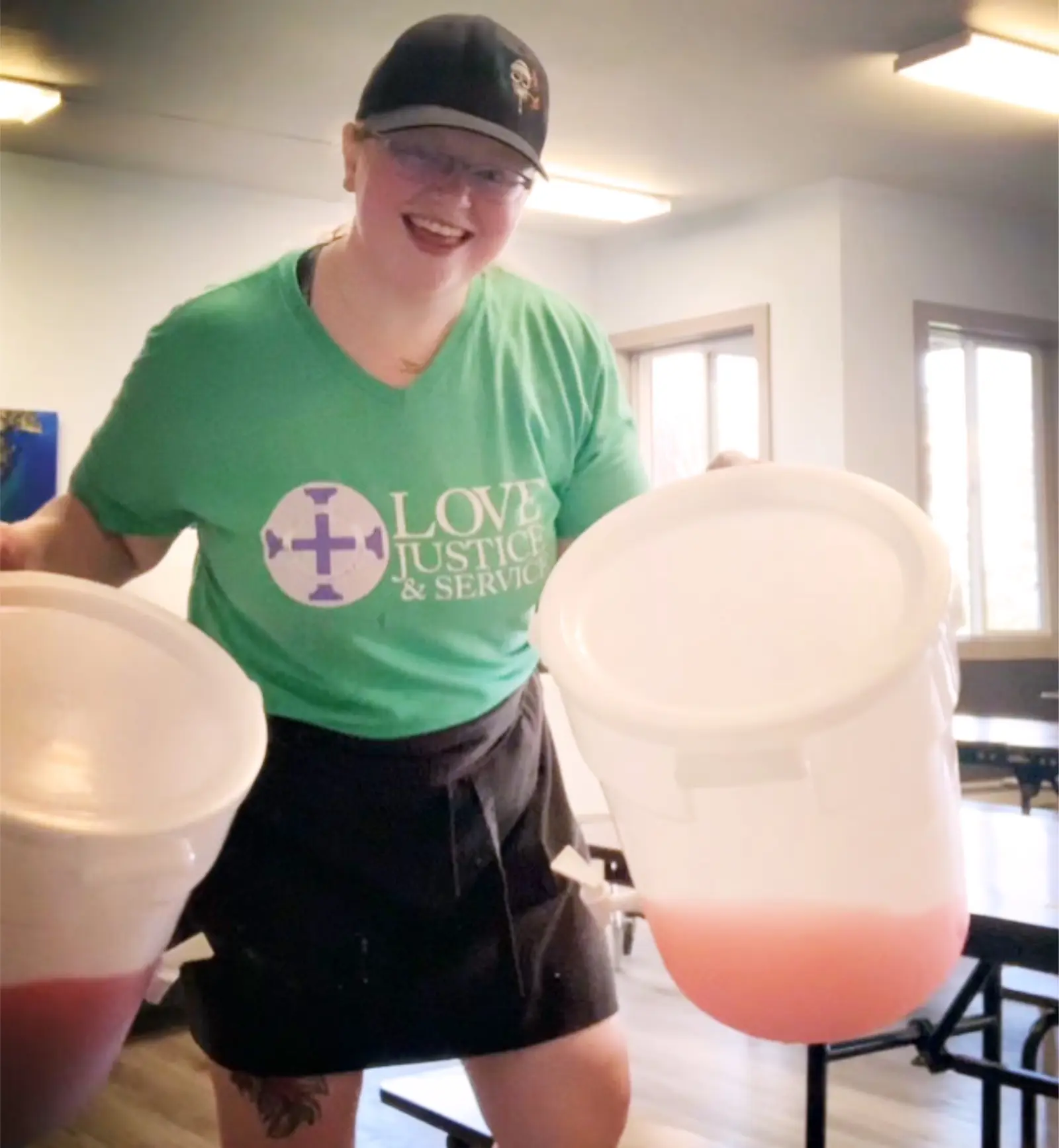 A smiling person in a green shirt and black apron holds two large white containers of pink liquid.