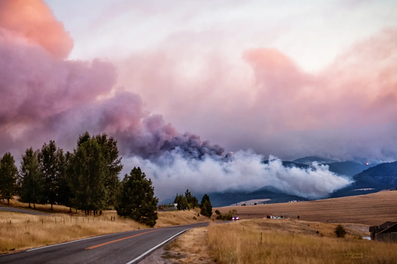 A road stretches through a landscape toward mountains beneath a smoke-filled, colorful sky.