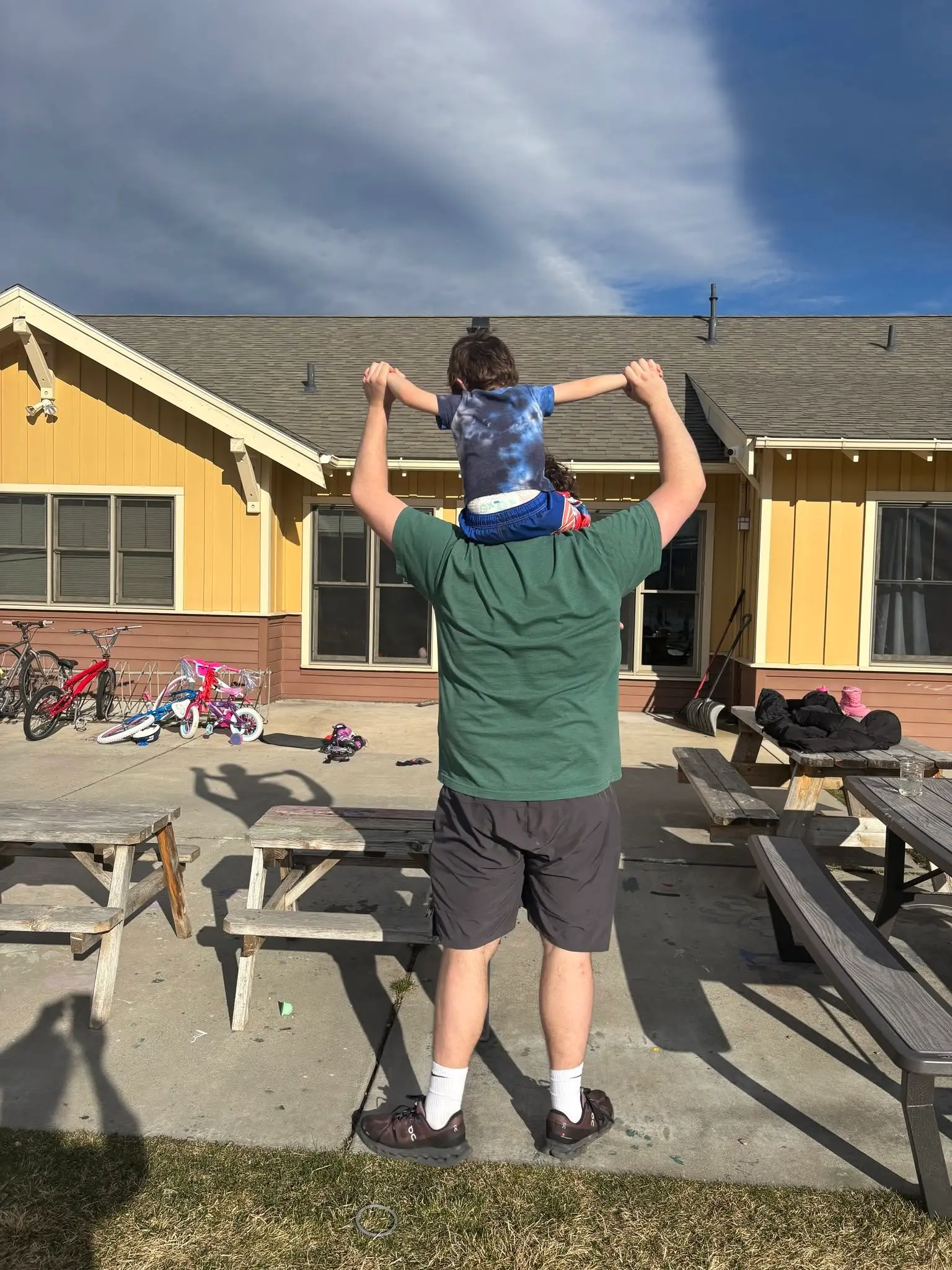 An adult carries a small child on their shoulders outside a yellow building.