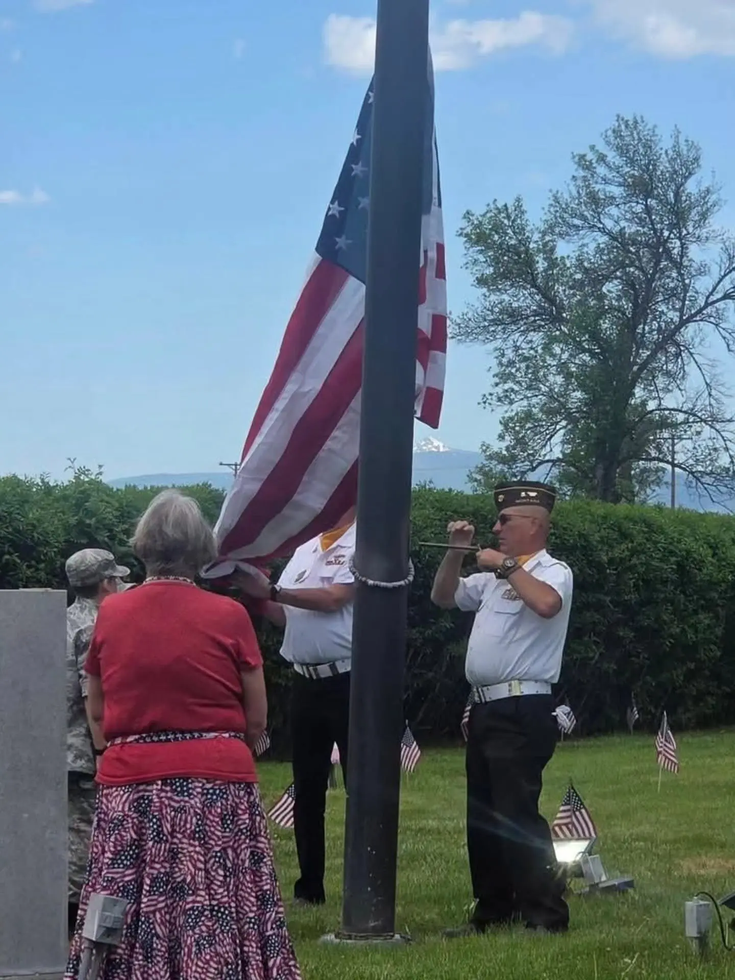 People gather outdoors to raise an American flag on a pole near a grassy area with small flags placed in the ground.