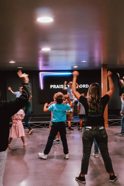 A group of children and an instructor are dancing in an indoor space with a screen displaying text in the background.