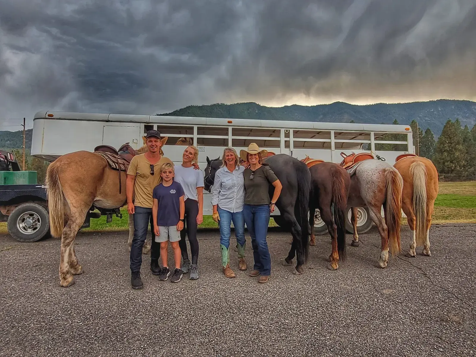 A group of people stand with horses in front of a horse trailer under a cloudy sky.