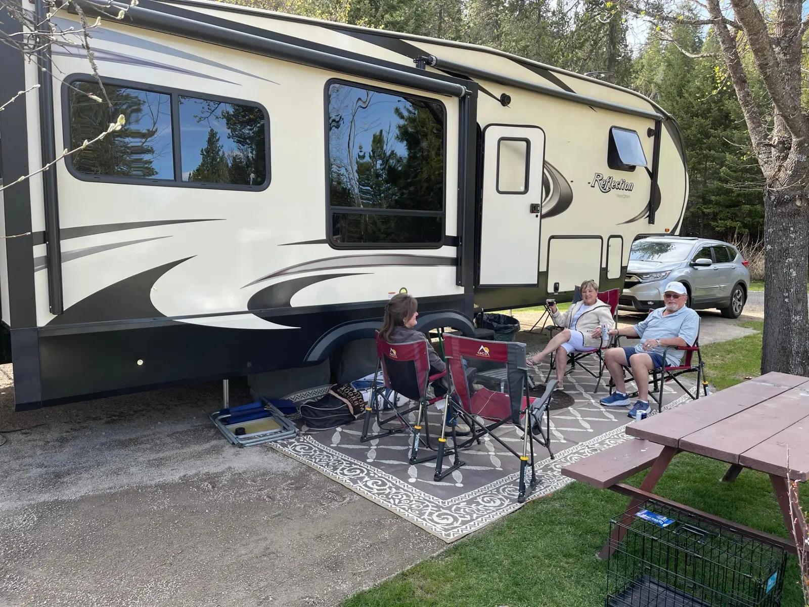 Fifth-wheel RV parked on a spacious wooded site with guests enjoying the outdoors