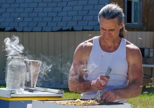 A man wearing a white tank top uses a tool while tending to a beehive with a smoker nearby.