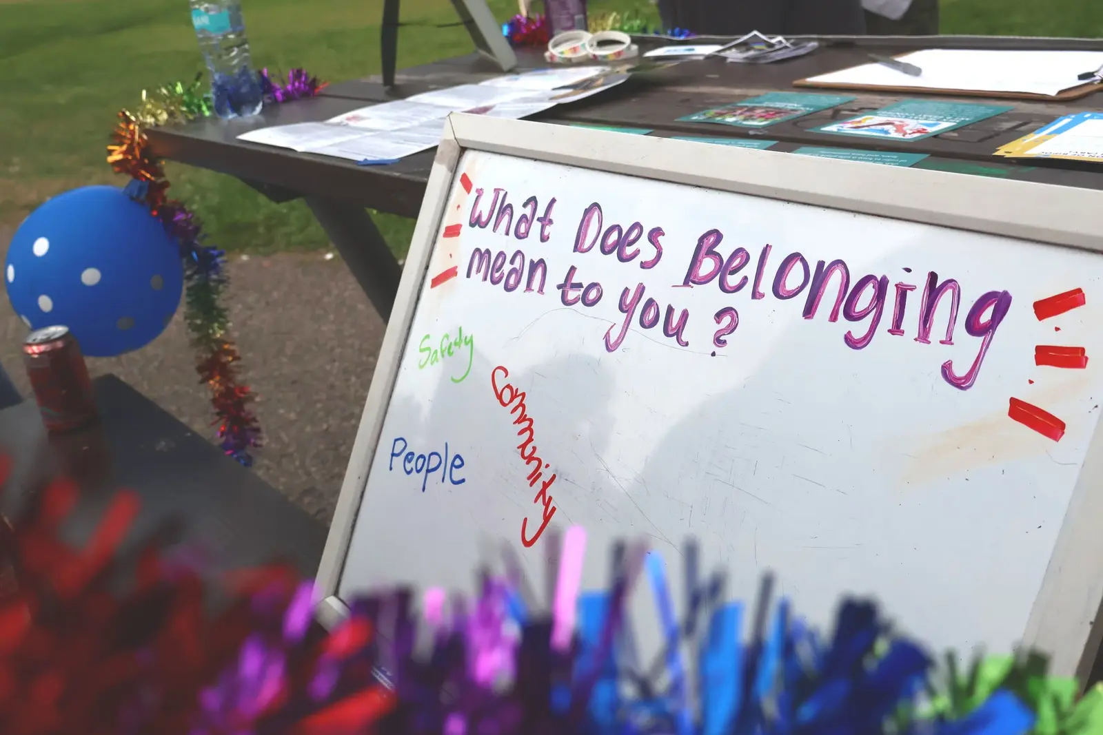 A whiteboard sits on a table outdoors with the question "What Does Belonging mean to you?" written on it in marker.