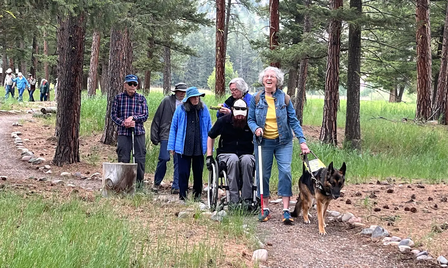 A group of adults, including one in a wheelchair and one with a service dog, walk along a forest trail.