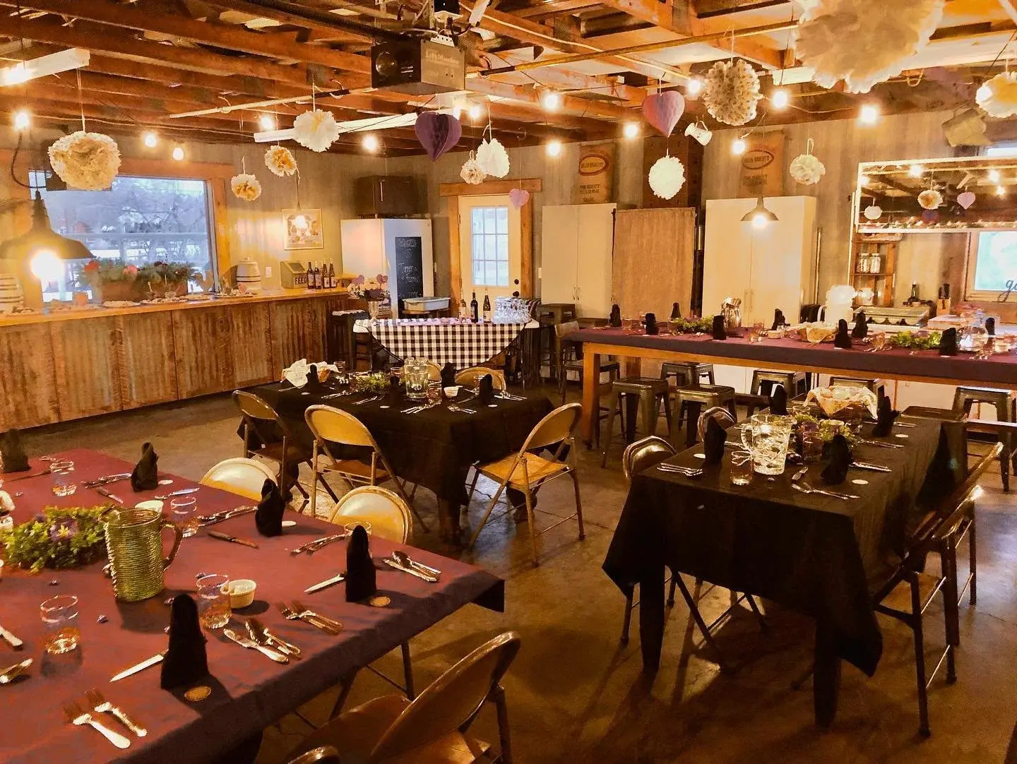 A rustic room is arranged with tables and chairs for a gathering, illuminated by festive string lights.