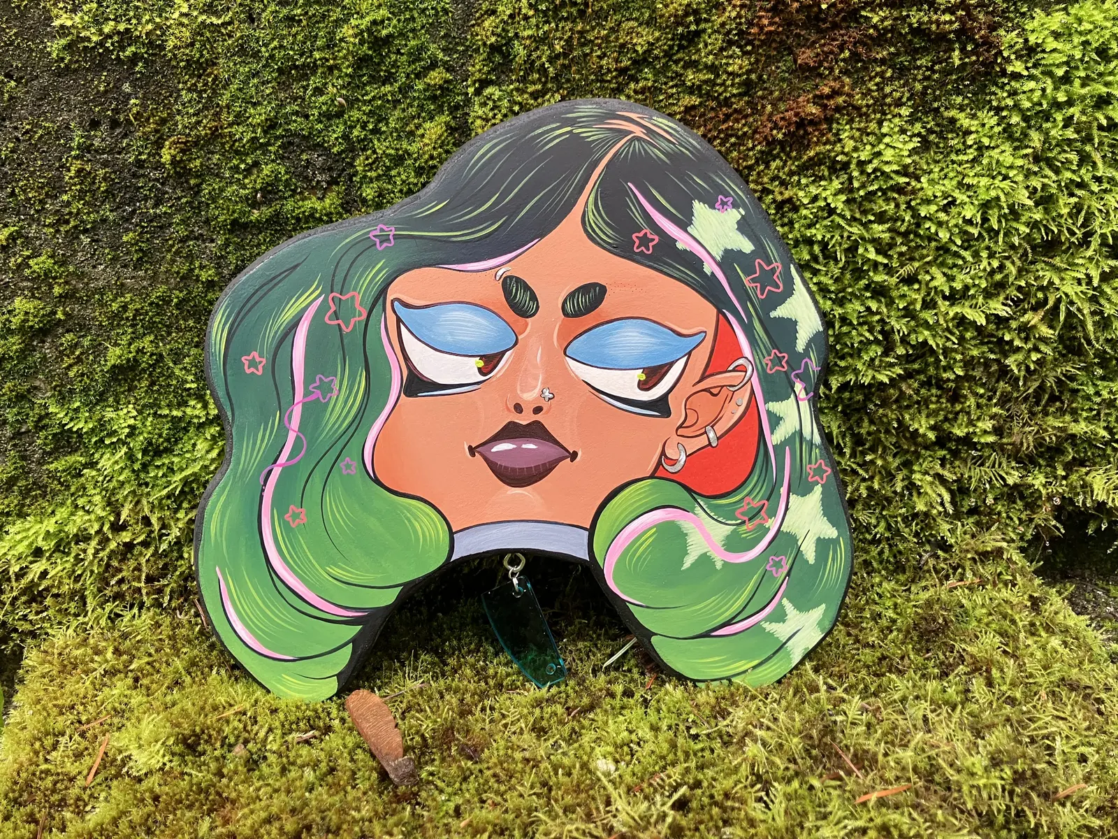 A colorful illustrated cutout of a woman's face with green hair rests on a mossy surface.