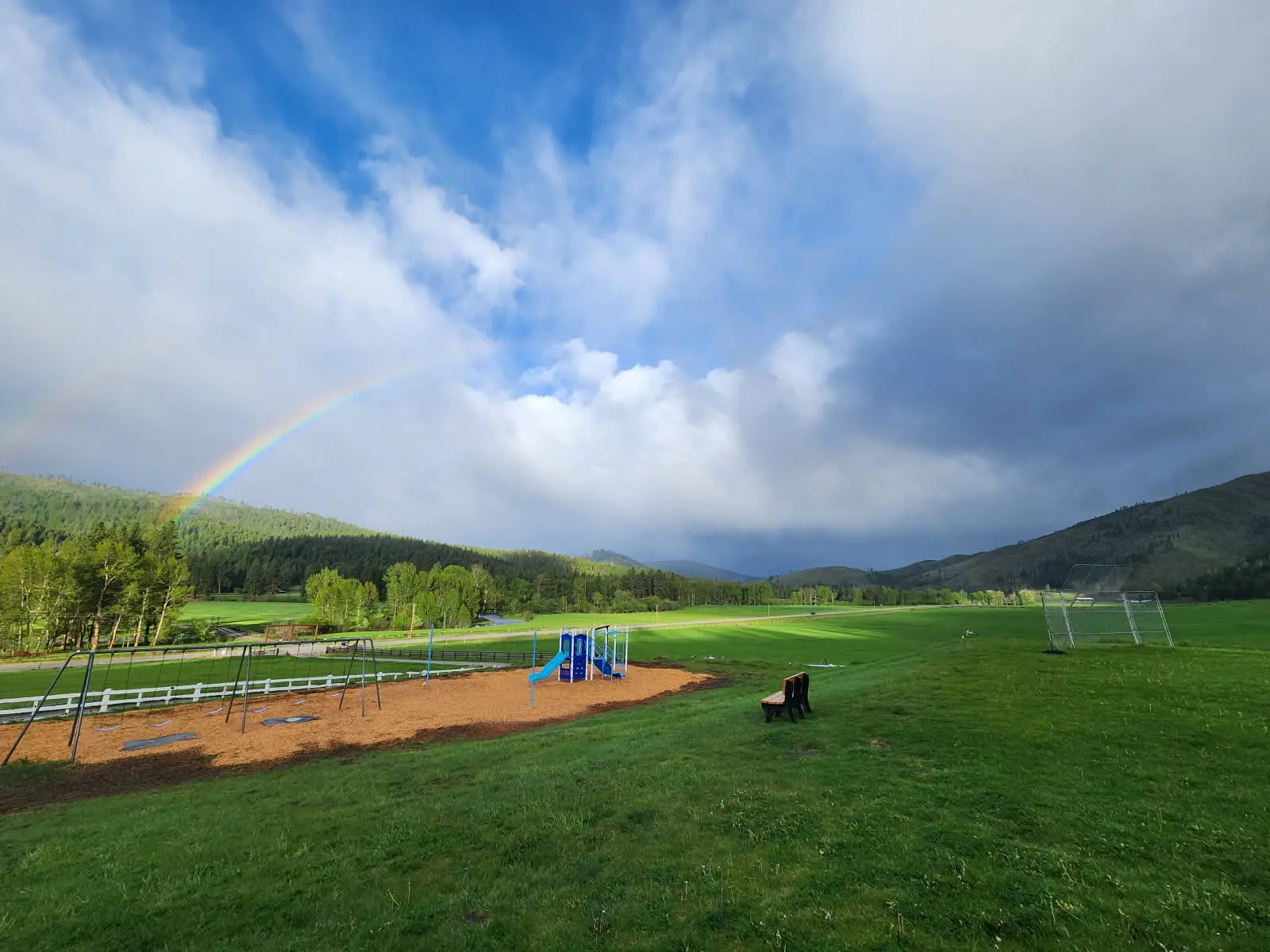 A rainbow arcs over a grassy field containing a playground and a park bench.