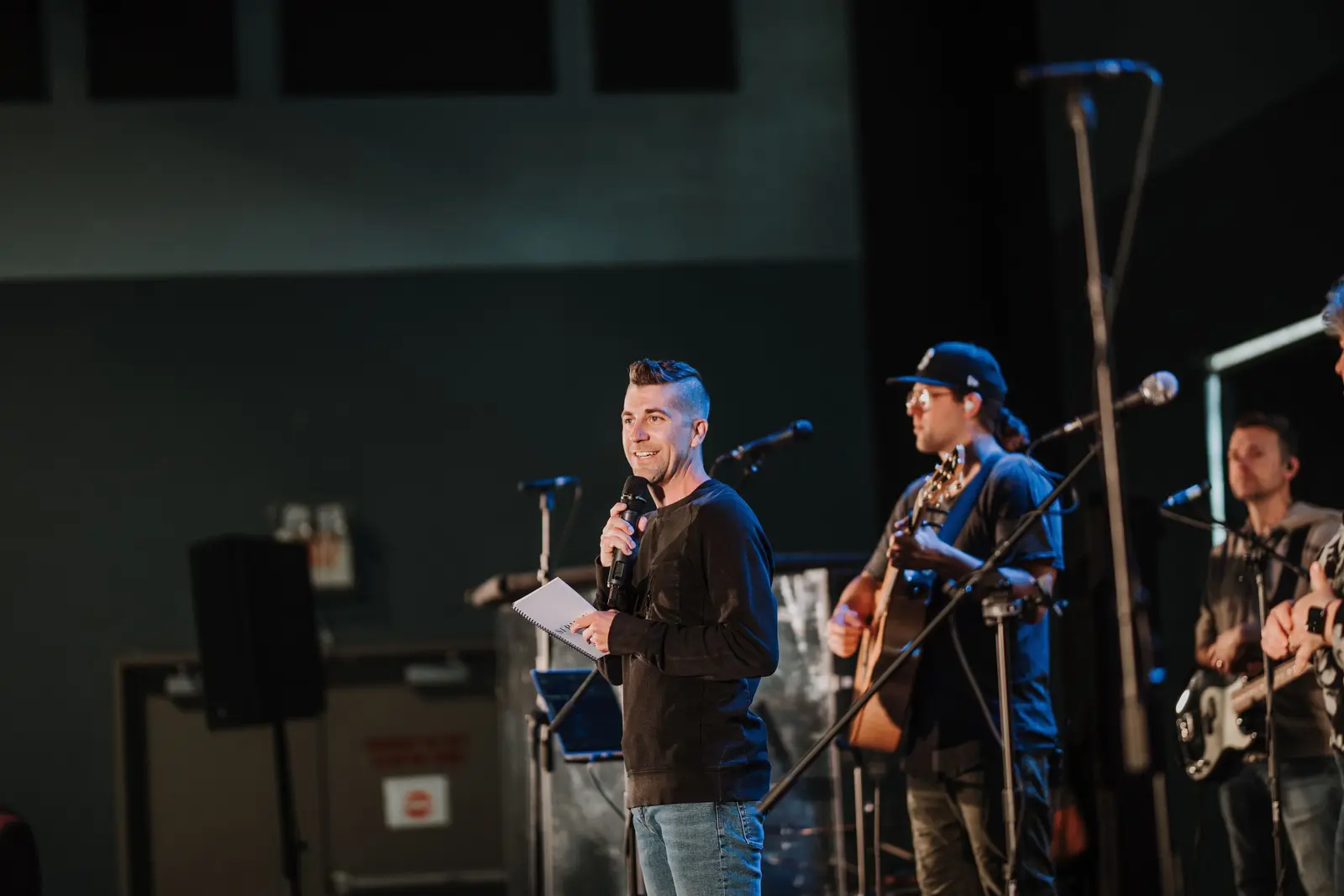 A man speaks into a microphone on stage while a band plays behind him.