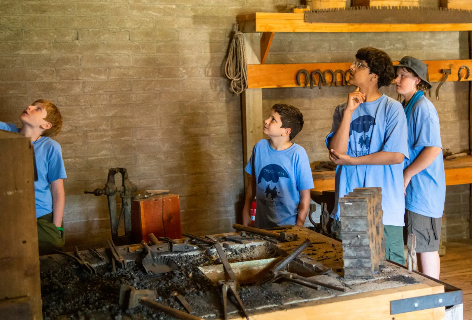 Four boys in matching blue t-shirts observe a blacksmith workshop display.