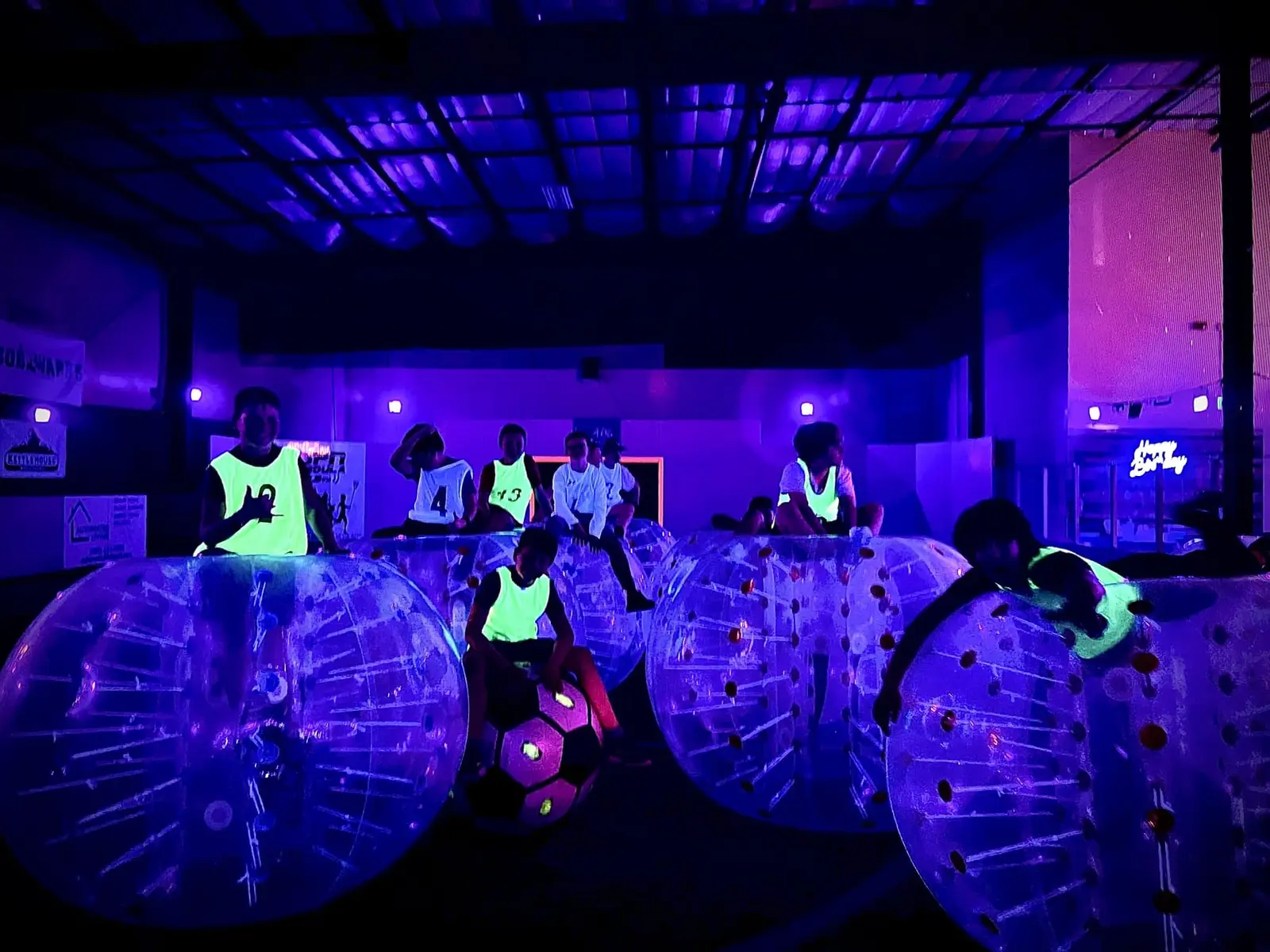 A group of people wearing neon vests inside bubble suits in a dark room illuminated by blacklight.