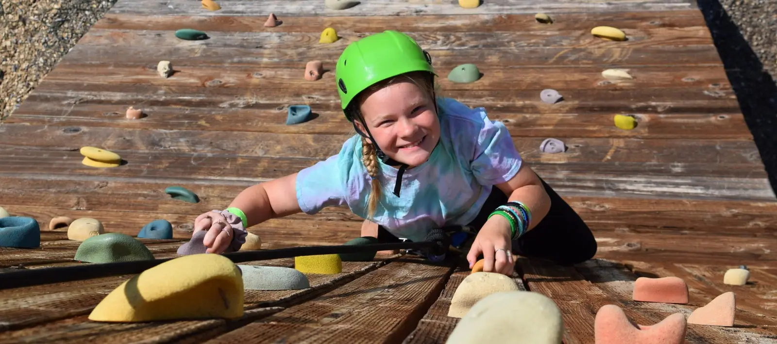 A child in a safety helmet climbs a wooden rock wall.