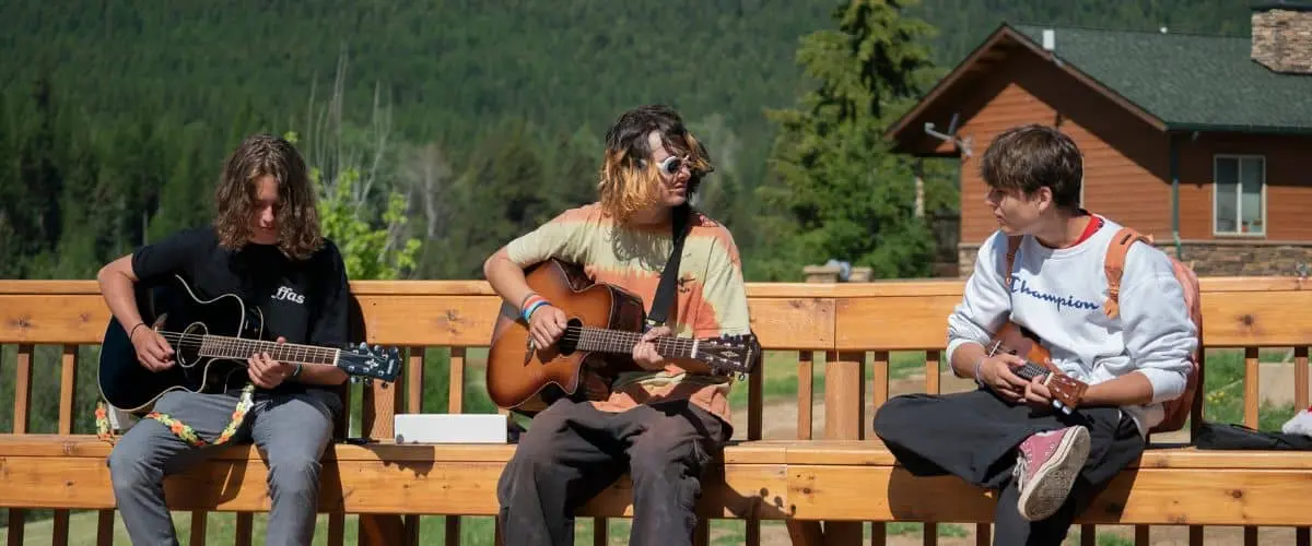 Three young people sit on a wooden bench playing stringed instruments outdoors.