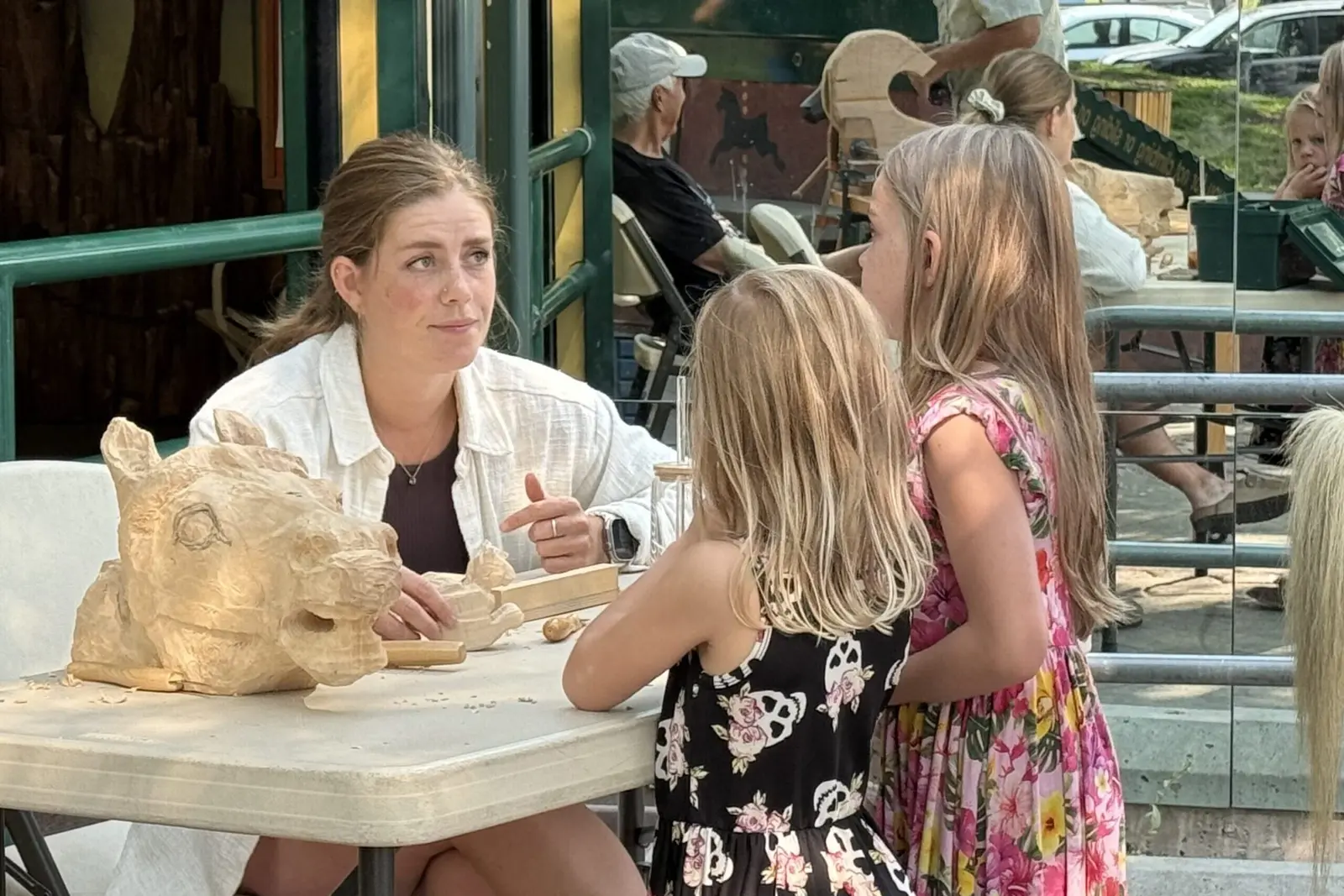 A woman instructs two young girls at a table with wood carvings.